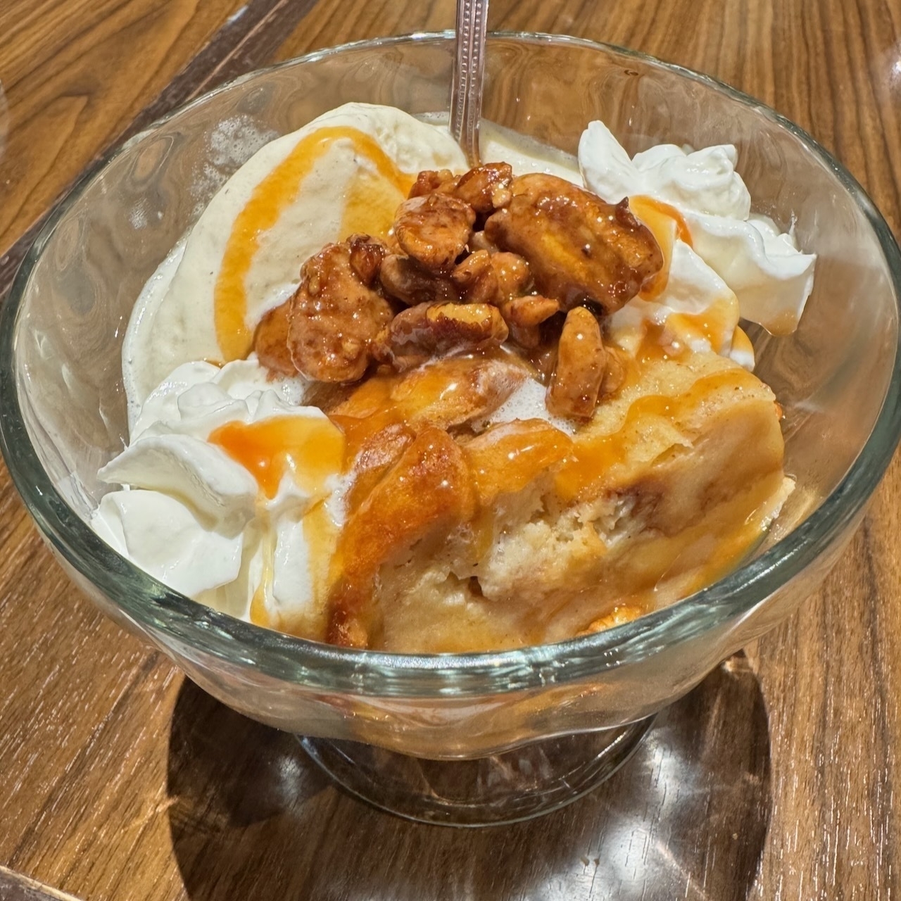 Auto-generated description: A dessert bowl containing ice cream, topped with whipped cream, caramel sauce, and candied nuts is placed on a wooden surface.
