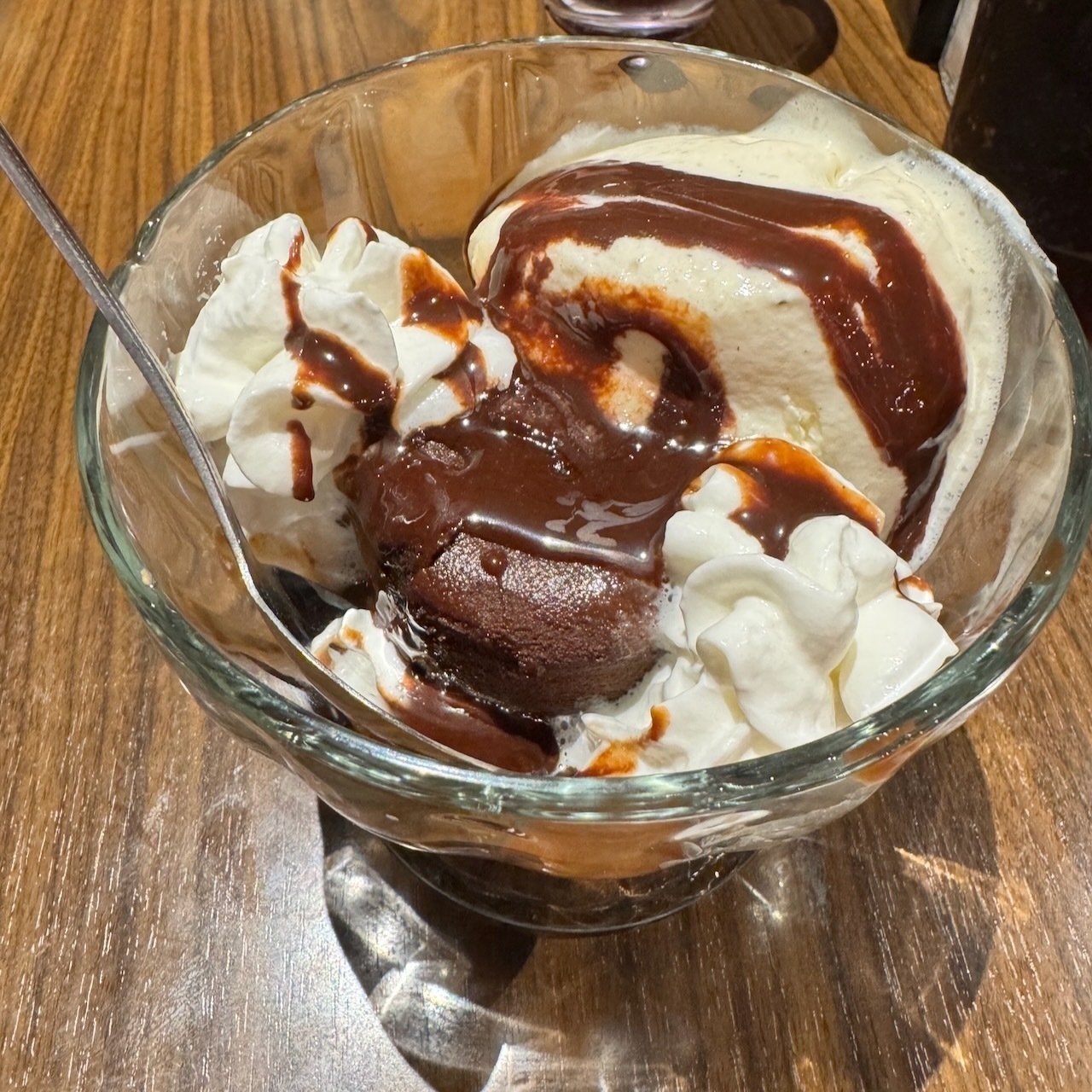 Auto-generated description: A glass bowl holds a dessert with vanilla ice cream, chocolate syrup, and whipped cream on a wooden table.