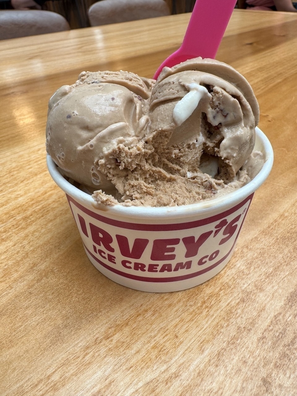 Auto-generated description: A scoop of ice cream in a branded cup is served with a pink spoon on a wooden table.