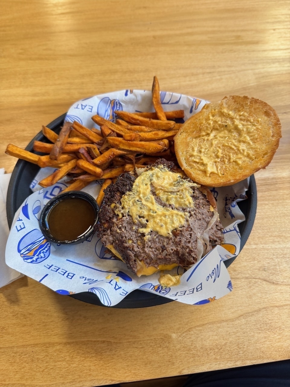 Auto-generated description: A burger with mustard on a bun, served with sweet potato fries and a small cup of sauce, is presented on a paper-lined plate.